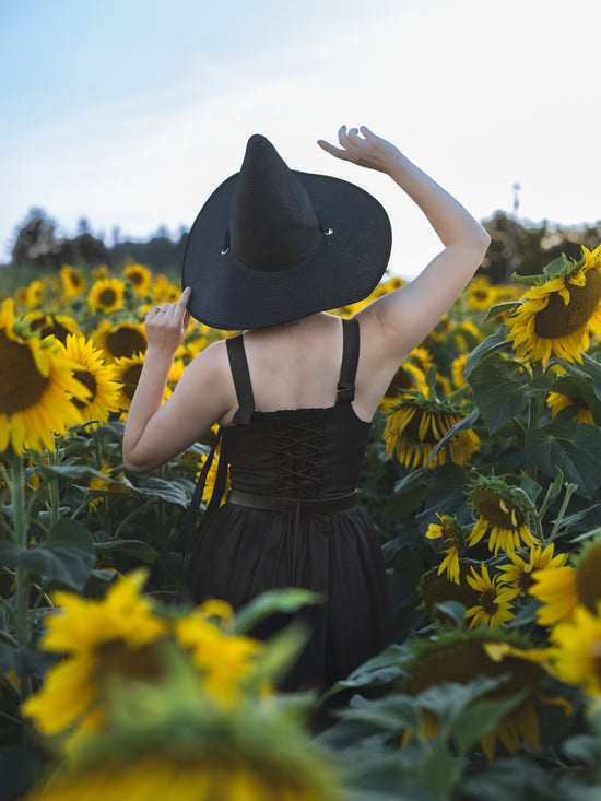 back view of model wearing witch hat and black lace up corset dress looking out into a field of sunflowers